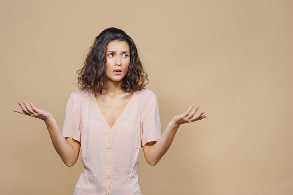 woman with wavy hair in a beige shirt on a beige background with her hands up is sad and indignant